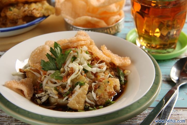 Kupat Tahu Magelang, a signature dish from Magelang, Central Java. It consists of rice cake (kupat or ketupat), bean curd (tahu), vegetable fritter, cabbage and beansprouts in sweet soy broth enriched with ground peanuts. Served on a white ceramic plate underlined with a green plate. A pair of spoon and fork is placed next to it. A tea in a glass is placed on the upper right hand side. An extra serving of crispy crackers and vegetable fritters are placed nearby. The meal is set on a rustic wooden table painted with bright colours of white and blue.